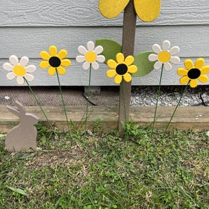 May include: Wooden garden decorations featuring daisy-shaped flowers with yellow and white petals and black centers. A wooden bunny silhouette is in the foreground. The flowers are on green stems, set against a gray background.
