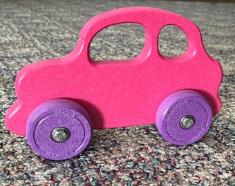 May include: A pink wooden toy car with purple wheels. The car has a simple design with rounded edges and a cutout for a window. The wheels are made of a textured material and have a silver screw in the center.