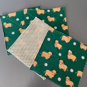 May include: Four green fabric pot holders with a white daisy pattern and a brown highland cow design. The pot holders are folded and stacked on top of each other.