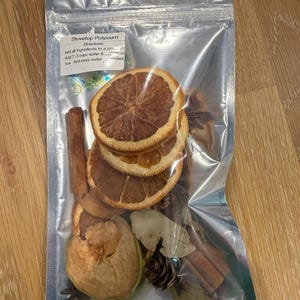 May include: A clear, sealed bag containing stovetop potpourri ingredients. The bag includes dried orange slices, cinnamon sticks, star anise, a dried apple slice, bay leaves, and a small pine cone. The label reads "Stovetop Potpourri".