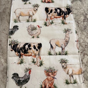 May include: White farm-themed kitchen towel featuring various farm animals, including cows, sheep, chickens, and pigs, each adorned with floral crowns. The animals are set against a white background with a subtle hexagonal pattern.