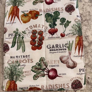 Puede incluir: Un paño de cocina cuadrado, blanquecino, con ilustraciones de diversas verduras como zanahorias, tomates, rábanos y ajo. El texto incluye "Heritage Carrots", "Sweet Tasty" y "Delicious Garlic".
