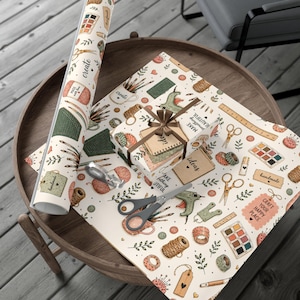 May include: A roll of wrapping paper and a gift box wrapped in the same paper, both featuring a repeating pattern of craft supplies including scissors, paint palettes, yarn, rulers, and glue guns, with the text "create," "ideas," and "handmade."