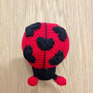 May include: A crocheted ladybug toy featuring a bright red rounded body with black heart-shaped spots. The head and lower appendages are crafted from solid black yarn, with two small red feet extending from the bottom of the dark base.