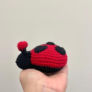 May include: A small, handcrafted crochet ladybug toy featuring a bright red body with black circular spots and a black head with two small red-tipped antennae. The stuffed insect is made from yarn with a visible textured stitch pattern throughout.