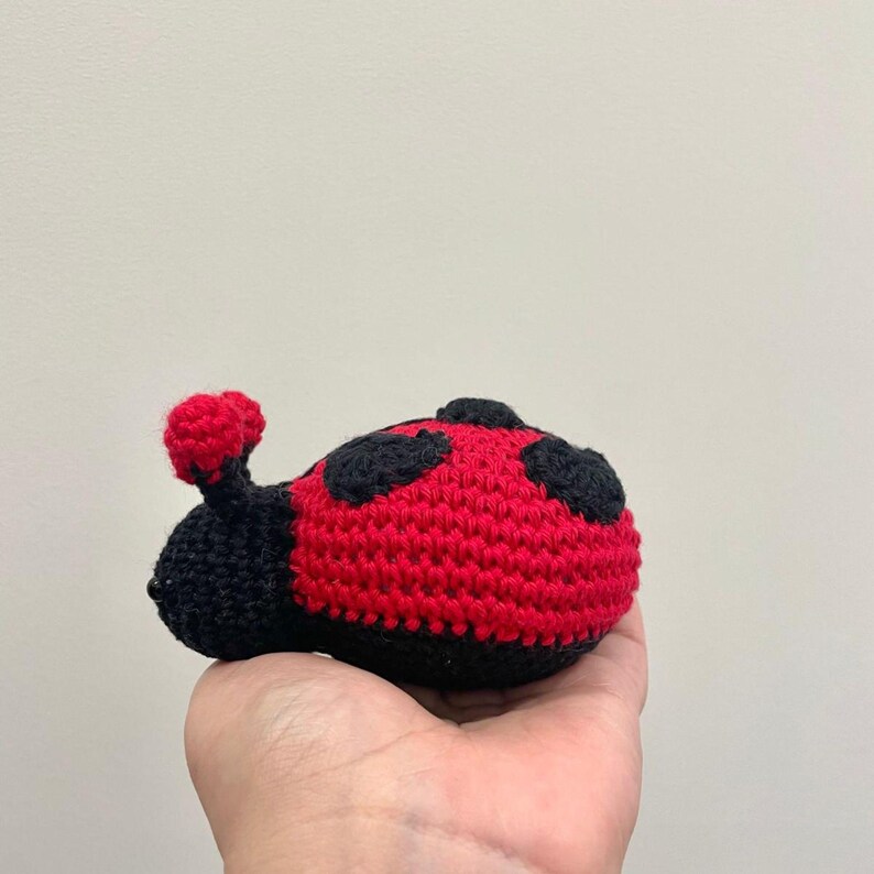 May include: A small, handcrafted crochet ladybug toy featuring a bright red body with black circular spots and a black head with two small red-tipped antennae. The stuffed insect is made from yarn with a visible textured stitch pattern throughout.