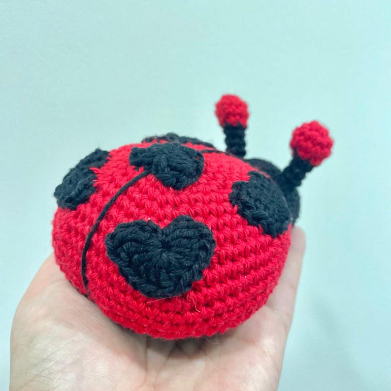 May include: A handcrafted crochet ladybug toy featuring a bright red rounded body adorned with black heart-shaped spots. The insect has two black antennae topped with small red spheres and a textured yarn surface, creating a soft, plush appearance.