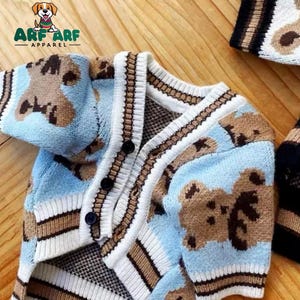 May include: A light blue knitted cardigan with brown teddy bear print. The cardigan has a button closure and white and brown stripes on the cuffs and hem.