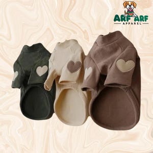 May include: Three dog sweaters in olive green, cream, and brown, each with a heart-shaped patch on the sleeve. The sweaters have a turtleneck design and are made of a soft material. The logo "ARF ARF APPAREL" is in the top right corner.
