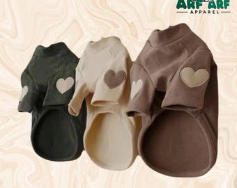 May include: Three dog sweaters in olive green, cream, and brown, each with a heart-shaped patch on the sleeve. The sweaters have a turtleneck design and are made of a soft material. The logo "ARF ARF APPAREL" is in the top right corner.