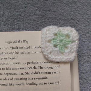 May include: A handmade, square crochet bookmark. The bookmark is white with a light green four-leaf clover design in the center. The bookmark is placed on the corner of a book. The book's text is visible.