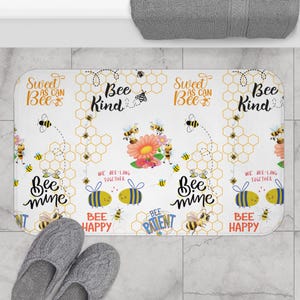 May include: A white rectangular bath mat with a bee-themed design. The mat features various phrases like "Bee Kind" and "Bee Happy" along with illustrations of bees, flowers, and honeycomb patterns. A pair of gray slippers is in the foreground.