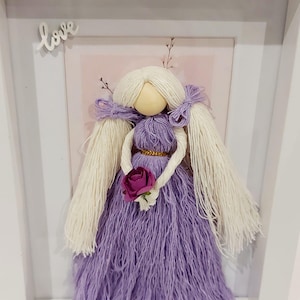 May include: A framed yarn doll with a wooden head, wearing a purple dress and holding a pink rose. The doll has long white yarn hair and is framed in a white shadow box. The word "love" is written on the frame.