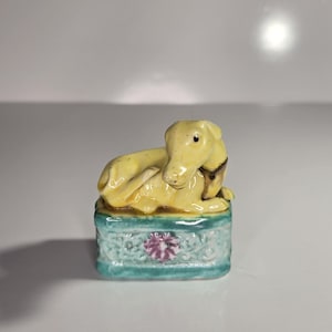 May include: A small, yellow ceramic dog figurine resting on a turquoise base with a floral design. The dog is lying down with its head up. The figurine is a decorative item.