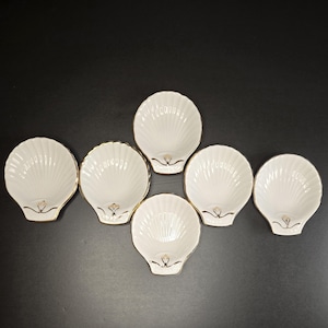 May include: A set of five white ceramic dishes shaped like scallop shells, each featuring a ribbed texture and a delicate gold-tone trim along the edges and a small gold-tone decorative accent near the base of the shell-shaped bowl.