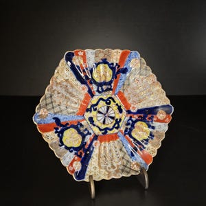 May include: A decorative ceramic plate with a scalloped edge, featuring a vibrant geometric and floral pattern in shades of cobalt blue, bright orange, yellow, and gold. The design is divided into segments with intricate gold detailing.