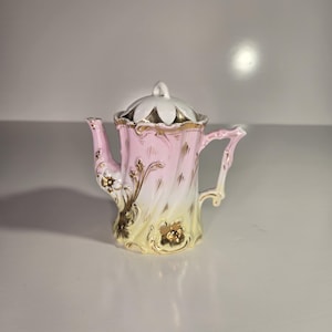 May include: A small, ornate pink and yellow porcelain teapot with a white lid. The teapot features gold accents and floral designs. The handle and spout are delicately curved. The lid has a flower-like design.