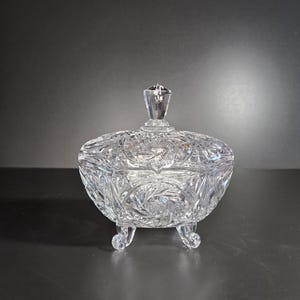 May include: A clear, cut-glass candy dish with a lid and three small feet. The dish features intricate floral and starburst patterns. The lid has a faceted knob. The dish is likely vintage and adds a touch of elegance.