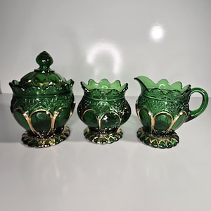 May include: A three-piece emerald green glass serving set. The set includes a lidded sugar bowl, a small open bowl, and a creamer with a handle. Each piece has a gold-colored trim and a decorative pattern.