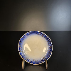 May include: A small, antique porcelain dish with a white center and a blue and gold decorative border. The rim has a scalloped edge. The dish is displayed on a small wooden stand against a black background.