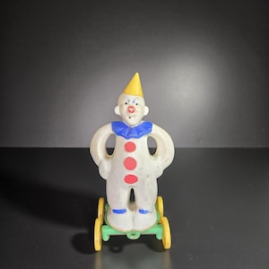 May include: A vintage toy clown figurine with a yellow cone hat, blue ruffled collar, and red button accents. The clown stands on a green platform with yellow wheels. The figurine has a white body and is posed with arms akimbo.