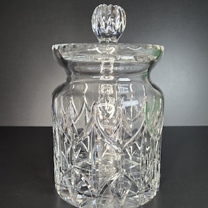 May include: A clear glass jar with a matching lid featuring a faceted diamond-cut pattern across the body. The lid has a round, textured knob handle. The glass is transparent and reflects light, showcasing the intricate geometric design.