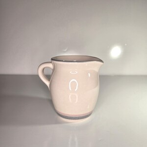 May include: A small, off-white ceramic creamer with a handle and a spout. The rim is outlined with a gray band, and a gray and pink band encircles the base. The creamer is designed for serving cream or milk.