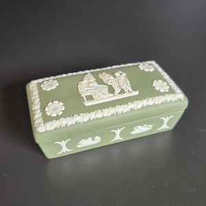 May include: A rectangular, sage green trinket box with white bas-relief floral and classical figures. The lid features a central scene of figures and a floral border. The box has a decorative floral pattern and X-shaped accents.