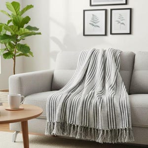 Puede incluir: Una manta a rayas grises y blancas con flecos está sobre un sofá gris claro. La manta tiene un tejido texturizado y un patrón de rayas verticales. Una mesa auxiliar de madera con una taza y una planta están en el fondo.