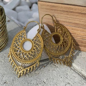 Peut inclure: Une paire de boucles d'oreilles créoles dorées avec des motifs filigranés complexes. Les boucles d'oreilles ont un design superposé et tombant avec un centre circulaire.