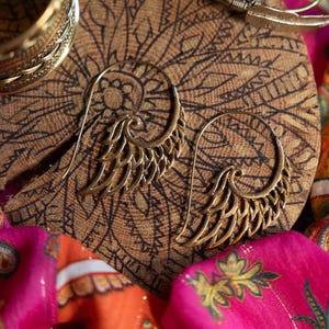 May include: A pair of gold-toned earrings with a wing design. The earrings are hanging on a wooden surface with a patterned design.