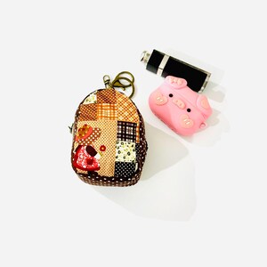 May include: A small brown patchwork fabric bag with a zipper closure and a key chain. The bag is shaped like a backpack and has a cartoon girl design. A pink pig-shaped silicone case is also in the image.