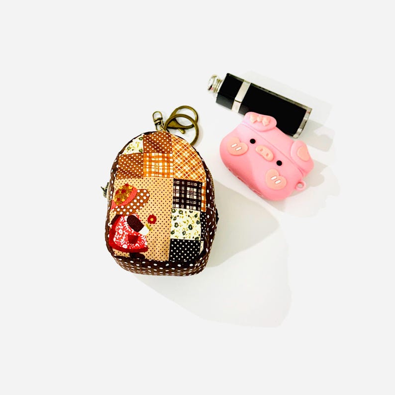 May include: A small brown patchwork fabric bag with a zipper closure and a key chain. The bag is shaped like a backpack and has a cartoon girl design. A pink pig-shaped silicone case is also in the image.