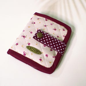 Peut inclure: Portefeuille matelassé à imprimé floral avec une bordure bordeaux et une sangle à pois. Le portefeuille est doté d'une fermeture à bouton et d'une plaque signalétique métallique qui indique "CHEZ VIES". Le fond est blanc.