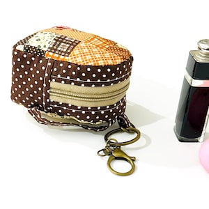 May include: A brown and beige patchwork fabric zippered pouch with a white polka dot pattern. The pouch has a brown strap and a gold key ring attached.