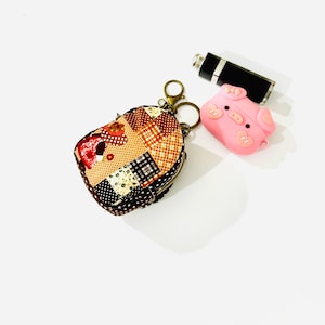 May include: A small patchwork fabric keychain with a zipper closure and a metal ring. The keychain is attached to a pink silicone pig keychain with a black nose and eyes.