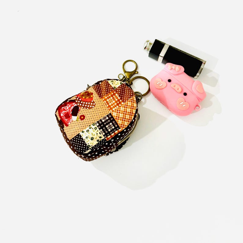 May include: A small patchwork fabric keychain with a zipper closure and a metal ring. The keychain is attached to a pink silicone pig keychain with a black nose and eyes.