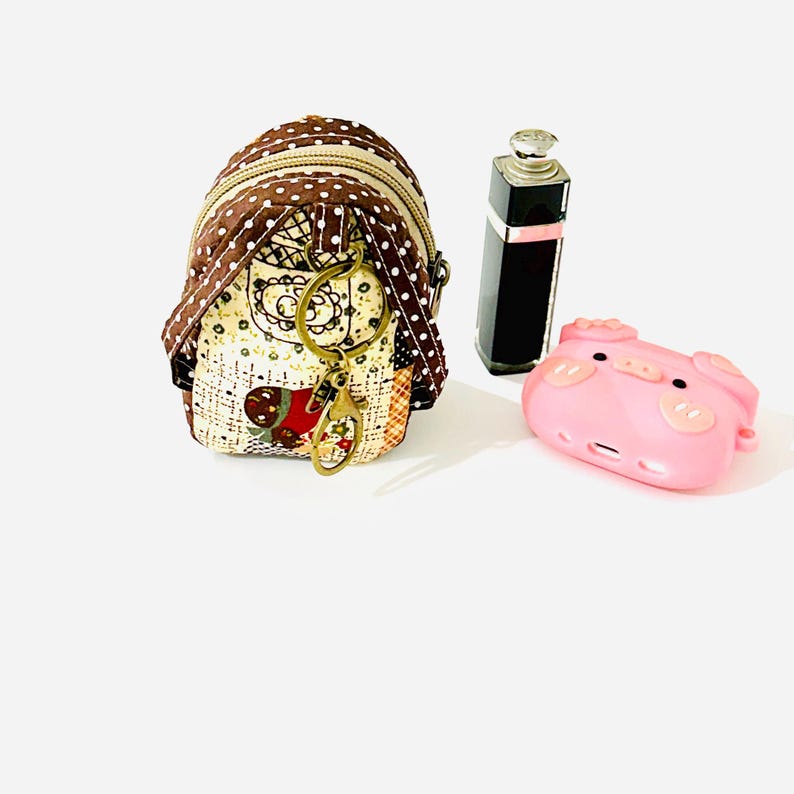 May include: A small brown and white polka dot backpack keychain with a gold key ring and a red and white heart design. The keychain is next to a pink pig-shaped phone case and a black bottle.