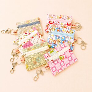 May include: A selection of small coin purses with floral patterns, zip closures, and keychains. The purses display diverse designs, including pink, blue, and yellow floral prints, and a patchwork design. Each purse is equipped with a silver-coloured keychain.