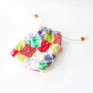 Könnte beinhalten: Eine kleine Patchwork-Kordeltasche mit einem roten, grünen, blauen und gelben Blumenmuster. Die Tasche hat einen weißen Kordelzug und Holzverschlüsse.