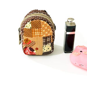May include: A small brown patchwork fabric backpack with a zipper closure and a brown strap. The backpack is decorated with a variety of floral and geometric patterns. A pink pig-shaped silicone phone case is also in the image.