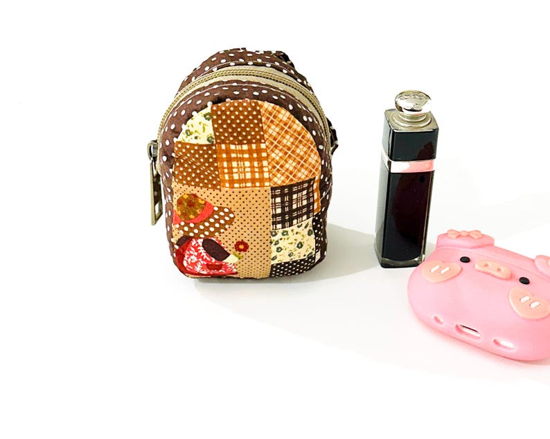 May include: A small brown patchwork fabric backpack with a zipper closure and a brown strap. The backpack is decorated with a variety of floral and geometric patterns. A pink pig-shaped silicone phone case is also in the image.