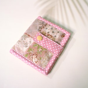 May include: A quilted fabric wallet featuring a floral pattern in shades of pink, beige, and green. The wallet is accented with pink and white polka dot fabric trim and a matching strap secured by a single yellow snap button closure.