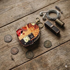 May include: A heart-shaped patchwork coin purse with a brass clasp, next to a black lipstick case, coins, and earbuds. The purse features a variety of patterned fabrics in shades of brown, red, and cream. The items are arranged on a rustic wooden surface.