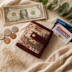 May include: A floral patterned wallet with a burgundy border and snap closure. The wallet has a small metal tag that reads "CHEZ VIES". US currency and coins are next to the wallet, along with credit cards.