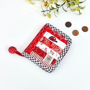 May include: A red and white patchwork fabric wallet with a black and white checked trim. The wallet has a zip closure and a small fabric loop.