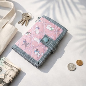 Könnte beinhalten: Ein rosa Stoffportemonnaie mit Hundemuster, das verschiedene Hundeillustrationen mit blauen und grünen Akzenten zeigt. Das Portemonnaie hat einen grauen Karoborder und einen hellblauen Knopfverschluss. Der Text "THE BEST" ist sichtbar.