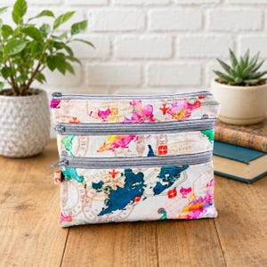 May include: A travel pouch with three zipped compartments. The pouch has a white background with a colourful map design, with pink, blue, and yellow watercolour-style splashes. The zips are grey.