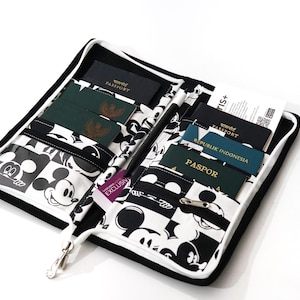 May include: Open passport holder with a black and white Mickey Mouse pattern. The interior has multiple pockets holding passports and travel documents. The holder is open, revealing a black zipper and a silver zipper pull.