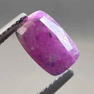 Puede incluir: Una gema rectangular facetada en tonos rosa y púrpura es sostenida por unas pinzas plateadas. La gema tiene una superficie pulida con inclusiones internas y un alto grado de transparencia, reflejando la luz.
